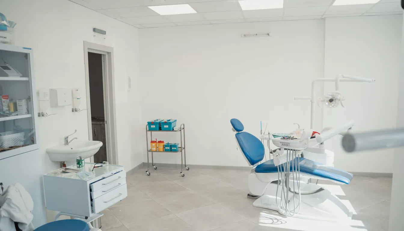 A bright, modern dental treatment room with a blue patient chair, sink and supply trolley.