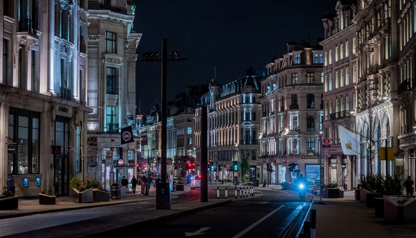 A wide London shopping street at night, lined with illuminated stone buildings and shopfronts, with a few pedestrians and a cyclist on the road.